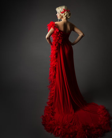 Fashion Woman in Red Luxury Dress with long Train Rear view. Elegant Lady in Silk Chic Gown with Feather Boa over Dark Gray Background. Blond Model Back with Bun Hair Updoの写真素材