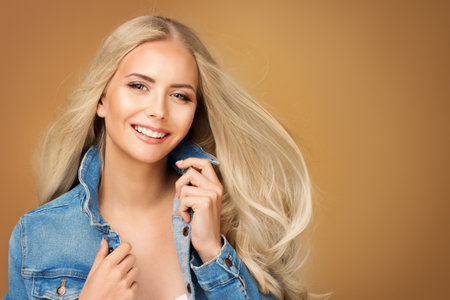 Happy Fashion Girl with Long Blond Hair. Beautiful Smiling Young Woman in Denim Jeans Jacket over Beige Background. Cheerful Model with Natural Make up and Summer Hairstyleの写真素材