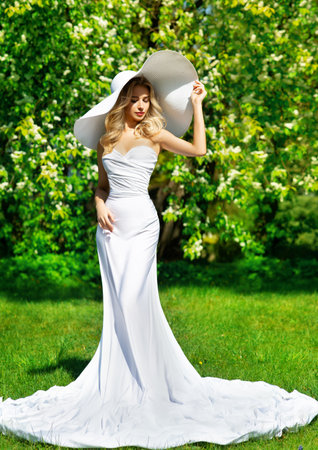 Woman in White Dress and Summer Hat outdoors. Beautiful Bride in Wedding Gown in Spring Blossom Park. Fashion Girl in luxury Clothes over Green Nature backgroundの写真素材
