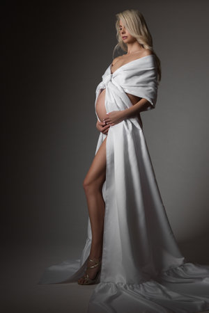 Pregnant Woman in White Dress looking at Belly. Beautiful Mother in Long Silk Gown over Dark Gray Background. Pregnancy Wedding Fashionの写真素材