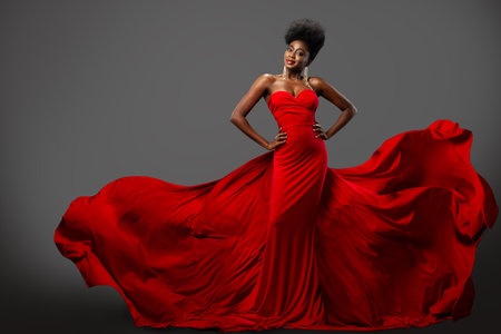 Fashion Woman in Red flying Dress with Afro Hairstyle. Dark Skinned Model in Silk Long Gown over Gray background. Happy elegant Lady wearing Luxury Clothesの写真素材
