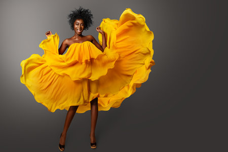 Happy smiling Woman in Orange Dress flowing on Wind over Gray. Fashion African Model with Afro Curly Hair in yellow Chiffon Gown. Cheerful Girl Carefree dancingの写真素材