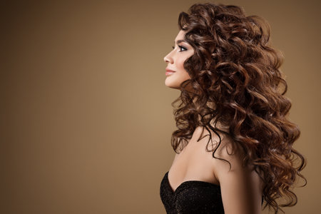 Beauty Model with Long Curly Hairstyle side view. Fashion Woman with Smooth Wavy Brown Hair. Beautiful Girl Face profile over beigeの写真素材