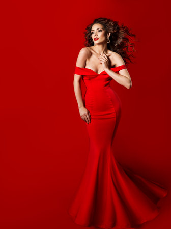 Elegant Woman in Long Red Dress. Fashion Model in Evening Silk Gown. Beautiful Girl with Curly Black Hairstyle waving on windの写真素材