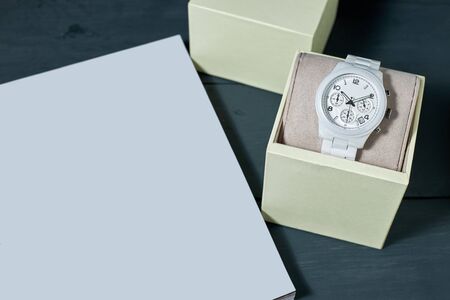 White ceramic wristwatch on a pillow in a beige box and white blank on a wooden background. Space for text.の写真素材