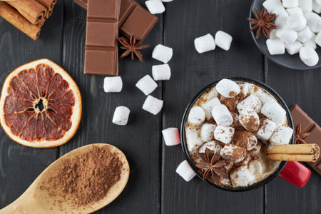 Red mug of hot chocolate with marshmallow, anise and cinnamon sprinkled with cocoa powder on a wooden table with slices of dried grapefruit and chocolate pieces. Winter hot drink. Top view.の写真素材