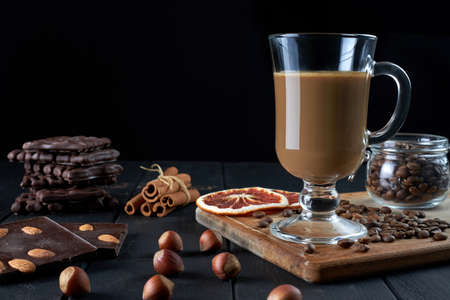 Glass of black coffee with milk with chocolate, hazelnuts, cinnamon sticks and slices of dried grapefruit on black background side view.の写真素材