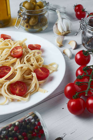 Delicious tagliatelle pasta with cherry tomatoes and basil on a white plate on a wooden table along with pasta ingredients: olives, tomatoes, garlic, olive oil and different types of dried peppers.の写真素材