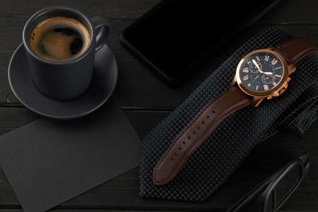 men's wrist watch on a black tie, a hot cup of coffee and a smartphone on a wooden table. black envelope with place for text.の写真素材