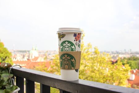 Prague, Czech Republic - May 2016: Starbucks paper cup of coffee stands on balcony railing on foreground. Beautiful golden autumn tree and bright city view on backgroundのeditorial素材