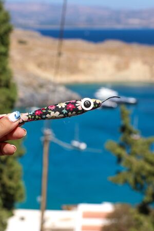 Soft toy colorful fish with a big eye, a button in a female hand with a manicure on the background of a bright Greek summer Rhodes beachの写真素材