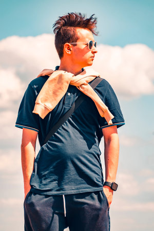 A handsome young man in dark glasses stands against the background of the sky and clouds and looks...
