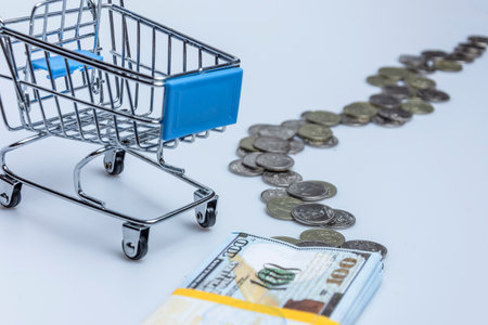 A supermarket trolley with a stack of hundred-dollar bills and scattered coins on a white background. The concept of online trading.の写真素材
