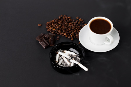 White cup of coffee with ashtray and coffee beans with chocolate on the black backgroundの写真素材