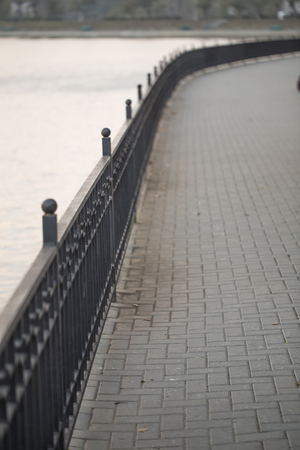 A paved walk and cast iron fence down the river bank. Tall trees in background, a beautiful perspectiveの写真素材