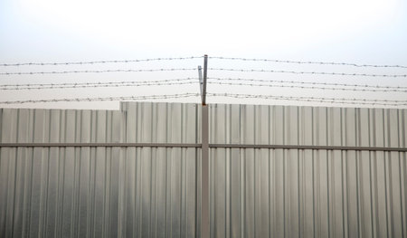 A closeup of perimeter fencing with barbed wire and construction fence against the sky