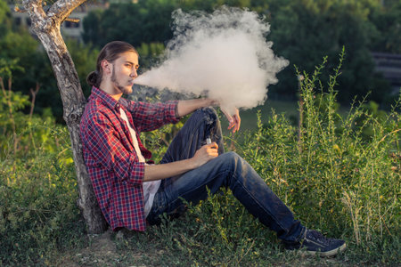 Handsome young man sitting on the grass and smoking electronic cigarette at outdoors.
