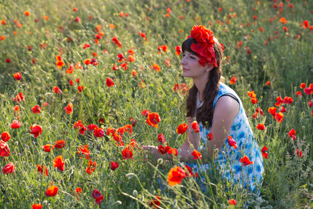 Beautiful romantic lady on the poppy field dressed in casual dress and floral wreath. Summer shot at sunset with backlight..の写真素材