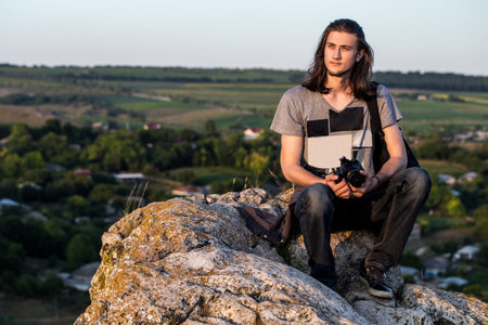 Handsome young longhair man sitting on a rocks with digital dslr camera against fields and blue...