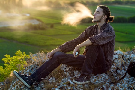 Handsome young man sitting on a rocks and smoking electronic cigarette at outdoors.の写真素材