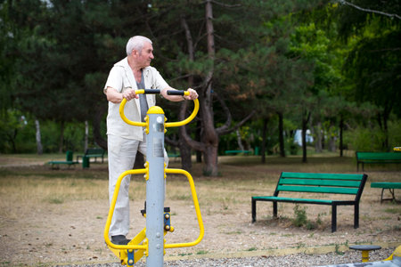 Old man making exercises on outdoor gym against green summer park as a background.
