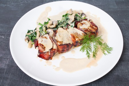 Bacon wrapped pork tenderloin with spinach and mushrooms. Plate located on a black table as a background.の写真素材