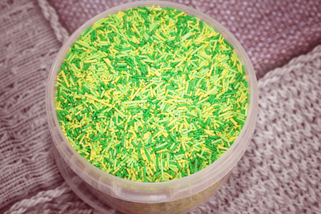 Sugar colourful sprinkles in a plastic pail. It is on a table covered with a patterned tablecloth. Close-up.の写真素材