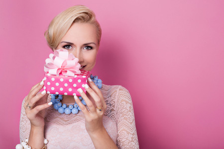 Happy birthday. Sweet blonde woman holding small gift box with ribbon. Soft colors. Studio portrait over pink background