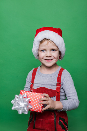 Smiling funny child in Santa red hat. Holding Christmas gift in hand. Christmas concept. Studio portrait over green backgroundの写真素材