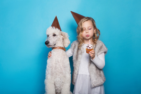 Little beautiful girl with dog celebrate birthday. Friendship. Love. Cake with candle. Studio portrait over blue backgroundの写真素材