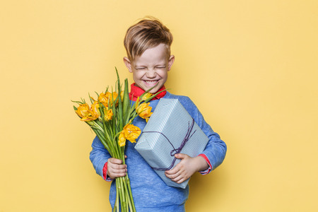 Handsome boy holding flower bunch and gift box. Valentine's day. Birthday. Mother's day. Studio portrait over yellow backgroundの写真素材