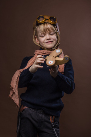 Happy child dressed in pilot hat and glasses. Kid playing with wooden toy airplane. Dream and freedom concept. Retro toned. Studio portrait over brown backgroundの写真素材