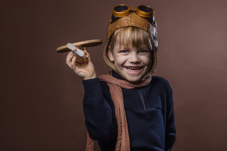 Happy child dressed in pilot hat and glasses. Kid playing with wooden toy airplane. Dream and freedom concept. Retro toned. Studio portrait over brown backgroundの写真素材