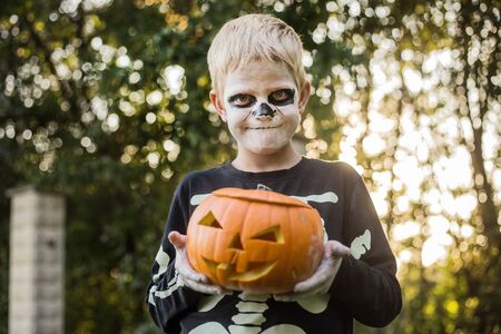 Happy young blond hair boy with skeleton costume holding jack o lantern. Halloween. Trick or treat. Outdoors portraitの写真素材
