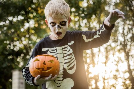 Happy young blond hair boy with skeleton costume holding jack o lantern. Halloween. Trick or treat. Outdoors portraitの写真素材