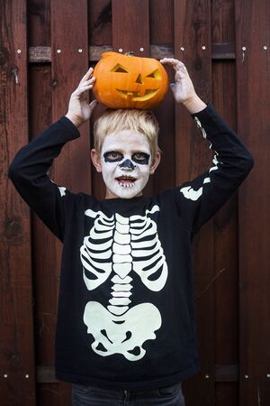 Happy young blond hair boy with skeleton costume holding jack o lantern. Halloween. Trick or treat. Outdoors portraitの写真素材
