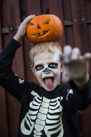 Happy young blond hair boy with skeleton costume holding jack o lantern. Halloween. Trick or treat. Outdoors portraitの写真素材