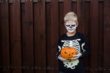 Happy young blond hair boy with skeleton costume holding jack o lantern. Halloween. Trick or treat. Outdoors portraitの写真素材