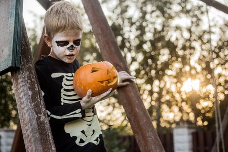 Happy young blond hair boy with skeleton costume holding jack o lantern. Halloween. Trick or treat. Outdoors portraitの写真素材