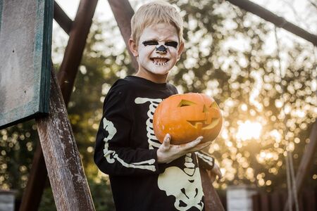 Happy young blond hair boy with skeleton costume holding jack o lantern. Halloween. Trick or treat. Outdoors portraitの写真素材