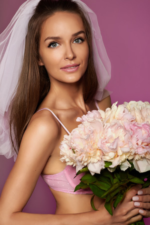 Pretty young sexy fiancee in white veil and pale pink lace lingerie with bouquet of peonies in handの写真素材