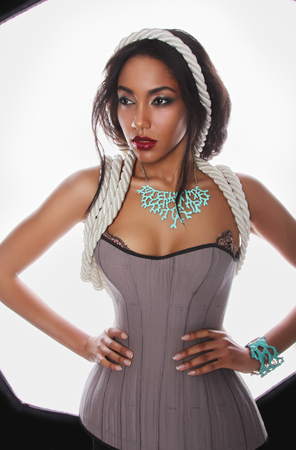 Fashion portrait of the beautiful mulatto whith dark skin and red lips in lingerie corset ropes and blue corral on her neck and hand on a white Backgroundの写真素材