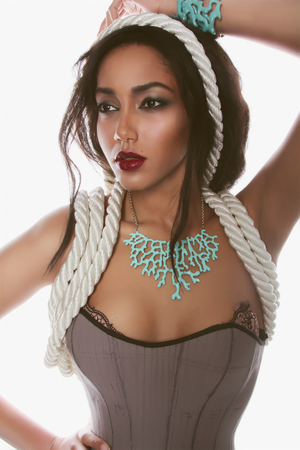 Fashion portrait of the Beautiful mulatto whith dark skin and red lips in lingerie corset ropes and blue corral on her neck and hand on a white Backgroundの写真素材