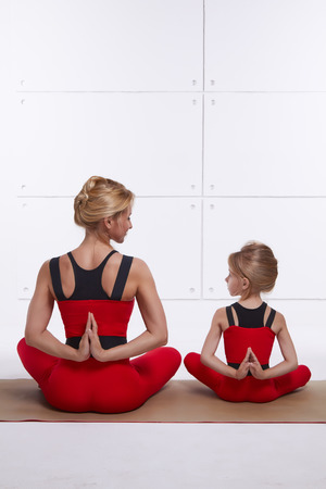 Mother and daughter doing yoga exercise, fitness, gym wearing the same comfortable tracksuits, family sports, sports paired siting back on relax pose and holding hand back they are on red skinny suit の写真素材