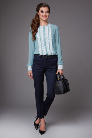 Beautiful sexy young business woman with evening make-up dressed in tight pants and silk blouse with lace long sleeves and high-heeled shoes and a small black handbag, business clothes for meetings and walksの写真素材