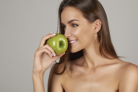 Young beautiful sexy girl with dark curly hair, bare shoulders and neck, holding big green apple to enjoy the taste and are dieting, healthy eating and organic foods, feeling temptation, smile, teethの写真素材