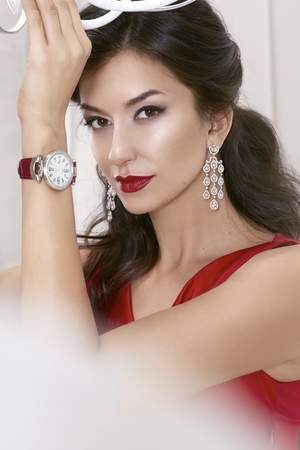 Beautiful sexy woman brunette Brown eyes in a red dress in lavish earrings with diamonds and watches on the Burgundy leather strap evening makeup, red lipstick, raised a hand upの写真素材