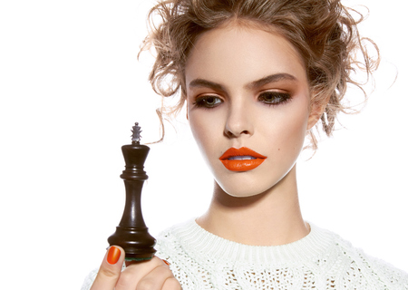 Beautiful young girl with a bright evening make-up, orange, scarlet lipstick, rosy cheeks, eye shadow, nail polish, blonde curly hair dressed in winter white knitted sweater holding a king chess pieceの写真素材