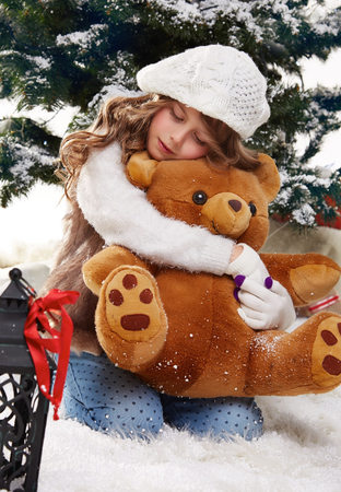 Beautiful little girl with long hair in a white winter sweater sits in the snow at Christmas trees and hugging a teddy bear gift for New Year lantern with a candleの写真素材