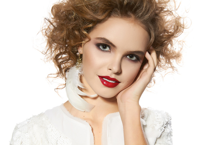 Portrait of beautiful young girl with a bright evening make-up perfect skin and blue eyes, red lips, nice smile blonde curly hair dressed in a white jacket with sequins on a white background in studioの写真素材